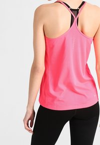 Donna che indossa un canottiera rosa brillante con schiena scoperta e leggings neri, vista di spalle su uno sfondo chiaro e semplice.