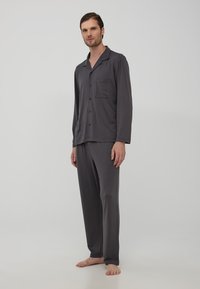 Pigiama grigio a maniche lunghe con camicia a bottoni dotata di taschino sul petto, abbinato a pantaloni larghi, realizzati in un tessuto morbido.