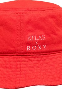 Rød bomuldsbucket hat med broderet tekst "ATLAS x ROXY" i hvid, en tekstureret stof og en let buet skygge.