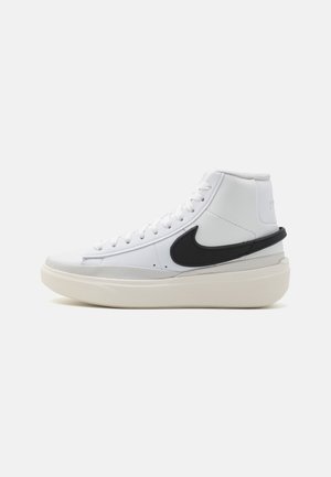 Białe wysokie sneakersy z grubą podeszwą, czarnym logo Nike po boku oraz białymi sznurówkami, pokazane z zewnętrznej strony.