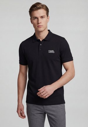 Ung mand iført en sort Karl Lagerfeld polo shirt og grå bukser, stående mod en ensfarvet lys baggrund.