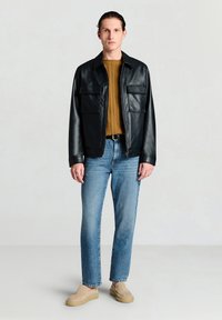 Schwarze Lederjacke mit zwei Fronttaschen, getragen über einem senffarbenen Pullover. Kombiniert mit hellblauen Jeans und beigen Slippern.