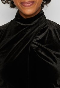 Personne souriante à la peau de teinte moyenne portant un haut en velours noir à col montant avec un détail de tissu drapé.