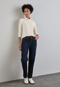 Haut crème à manches longues avec un col haut, associé à un jean en denim bleu foncé. La mannequin se tient contre un fond gris, portant des baskets blanches avec des accents.