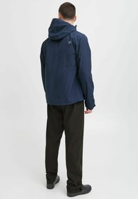 Jachetă impermeabilă de marină cu glugă ajustabilă, închidere cu fermoar, manșoane elastice și textură simplă, purtată cu pantaloni negri.