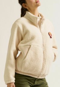 Cremefarbene Fleecejacke mit hohem Kragen, Reißverschluss, zwei Fronttaschen und einem runden Patch, der "Mom" in orange- und rötlichen Tönen zeigt.