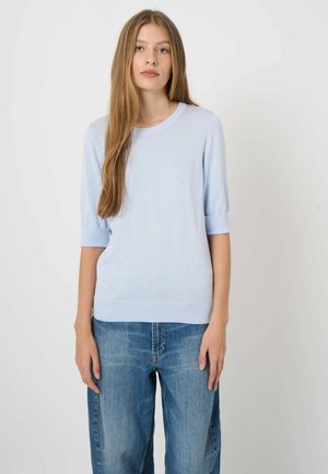 Junge Frau mit langen, glatten Haaren, die einen hellblauen Kurzarmpullover und lockere blaue Jeans trägt, vor einem schlichten weißen Hintergrund stehend.