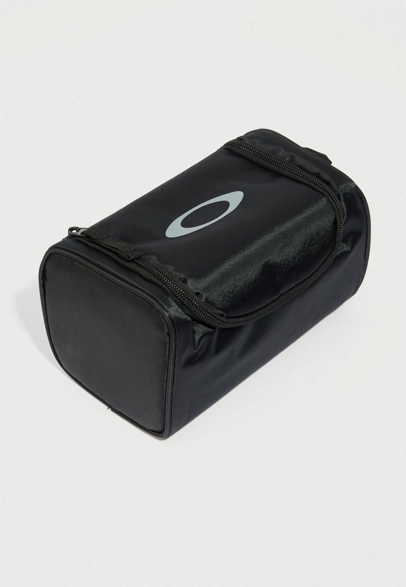 Pochette rectangulaire noire zippée avec un logo ovale gris sur le dessus, en tissu texturé, posée sur un fond clair uni.