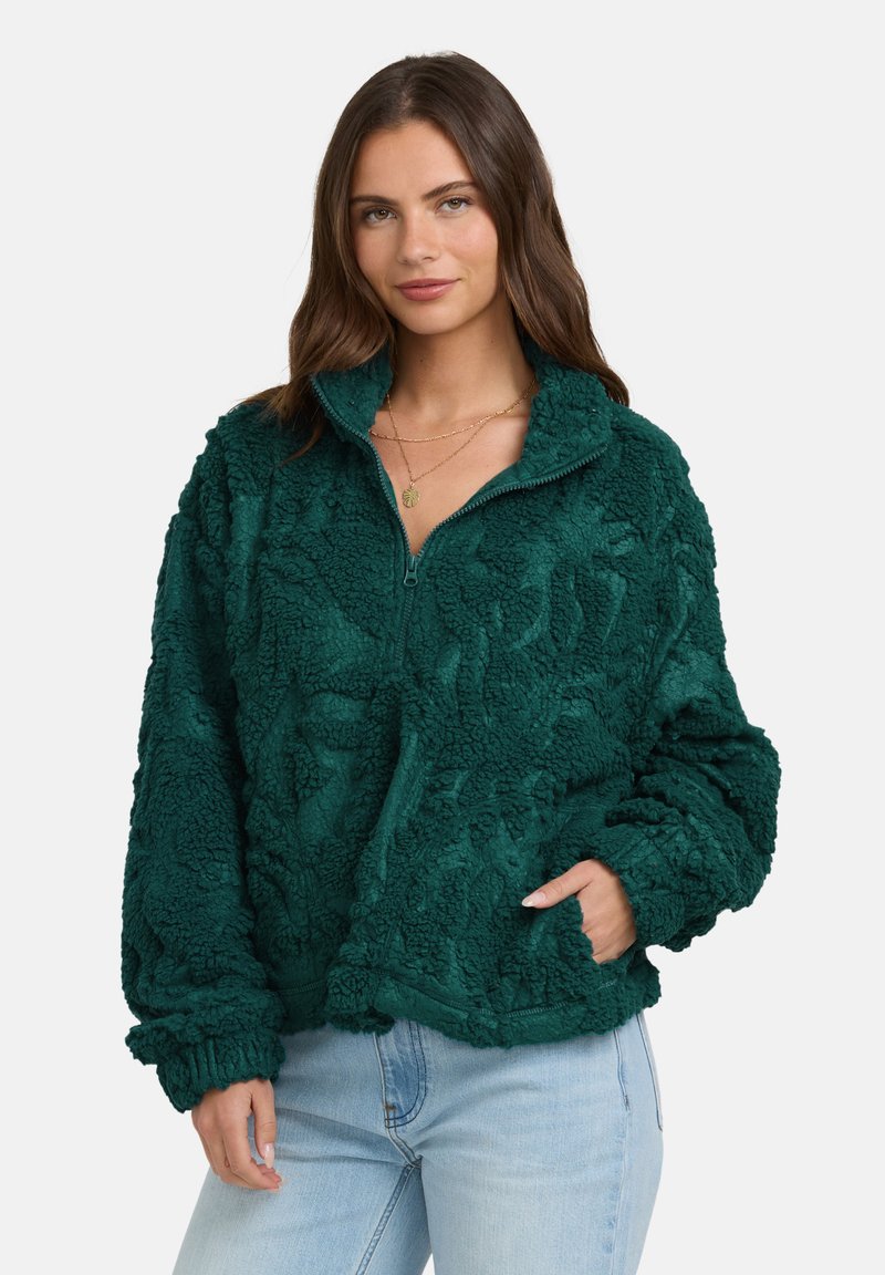 Eine Frau trägt eine taillierte, strukturierte Fleece-Halbzip-Jacke in Türkis mit langen Ärmeln und hellblauen Jeans und posiert mit einer Hand in der Jackentasche.