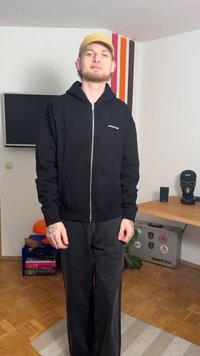 Melnā zip-up džemperis ar priekšējām kabatām, teksturēts audums, kopā ar vaļīgām melnām biksēm. Fonā redzams koka galds un dekoratīvi priekšmeti.