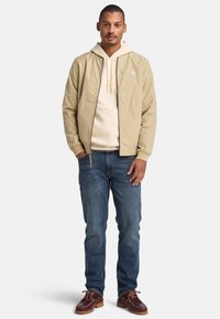 Chaqueta beige con cremallera sobre una sudadera clara, combinada con jeans azules y zapatos marrón oscuro. La chaqueta tiene puños elásticos y un detalle de logo.