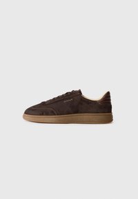 Brown suede sneaker with lace-up closure, tan rubber sole, and subtle branding on side. Features a streamlined silhouette and textured accents.