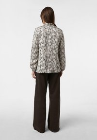 Woman with long brown hair wearing a long-sleeve snakeskin print blouse and dark wide-leg pants, standing facing away on a plain background.