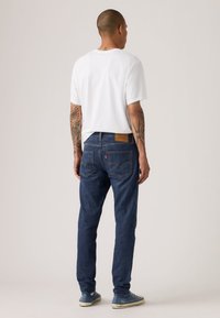 Σκούρο μπλε τζιν με στενή εφαρμογή, πέντε τσέπες και καφέ δερμάτινη ετικέτα στη ζώνη του πίσω μέρους. Φοριέται με λευκή μπλούζα και μπλε αθλητικά παπούτσια.