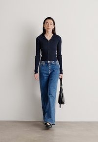 Jūras pielietojuma ribots zip-up tops, augsto jostasvietu gaismas zilo platu biksu komplekts, melna rokassoma un melnas kurpes uz neitrāla fona.