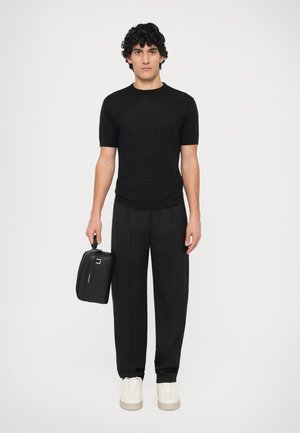 Camisa de punto negra con textura y de manga corta, combinada con pantalones anchos negros y zapatillas blancas. Sosteniendo un bolso rectangular negro con un asa.