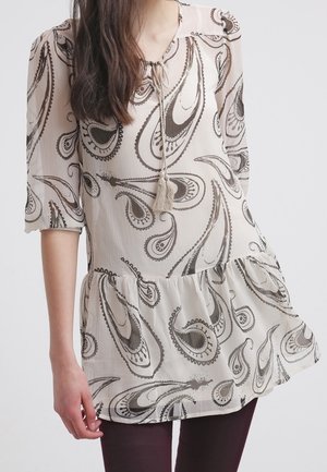 Kobieta ubrana w kremową tunikę z czarnymi wzorami paisley oraz rękawami 3/4, połączoną z ciemnymi dopasowanymi spodniami.