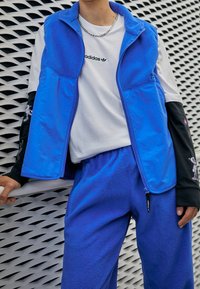 Blå fleeceväst med dragkedja, ihopparad med matchande byxor. En vit t-shirt under med tryckt logotyp. Svarta ärmar med grafiska detaljer.