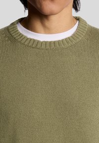 Pull en maille vert olive avec un col ras du cou côtelé et une texture lisse, comportant une couche blanche en dessous. Vue en gros plan des détails du tissu.