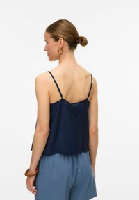 Navy blauwe mouwloze top van textuurstof, met verstelbare bandjes en een ontspannen pasvorm, gecombineerd met lichtblauwe shorts en een houten accessoire.