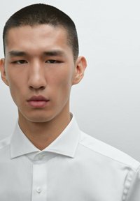 Weißes Button-Down-Hemd mit strukturiertem Kragen und vorderer Leiste. Das Modell hat kurzes, dunkles Haar und einen neutralen Gesichtsausdruck vor einem hellen Hintergrund.