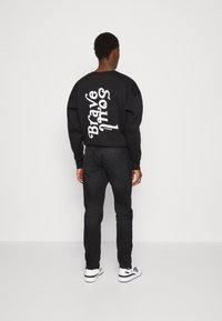 Hombre de espaldas vestido con una sudadera negra con el texto "Brave Souls London" en blanco, jeans negros y zapatillas blancas, contra un fondo simple.