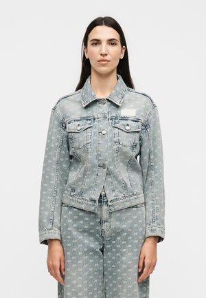 Femme aux longs cheveux foncés portant une veste en denim bleu clair et un jean assorti avec un motif répétitif de logo blanc sur un fond uni.