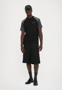 Man wearing black and green short-sleeve shirt, black shorts, gray socks, black slip-on shoes, and a black leather baseball cap standing.