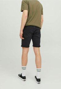 Joven vistiendo una camiseta verde oliva, pantalones cortos de mezclilla negros con dobladillo enrollado, calcetines blancos con rayas grises y zapatillas de lona negras, de pie mirando hacia atrás.