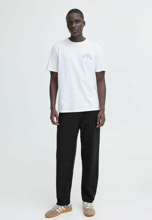 Weißes Baumwoll-T-Shirt mit kurzen Ärmeln und minimalistischem Grafikdruck, kombiniert mit schwarzen Hosen und grauen Sneakern mit weißen Akzenten.