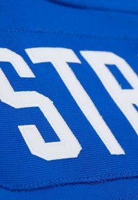 Gros plan sur l'inscription blanche cousue "STA" sur un tissu en bleu côtelé, montrant la texture détaillée et les bords de la broderie.