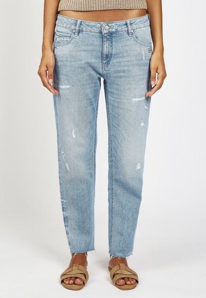 Model wearing light blue distressed jeans with frayed hems and beige cross-strap flat sandals, standing against a plain background.