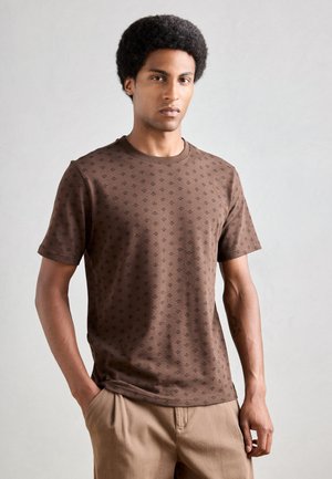 Brun t-skjorte med kort erme som har et subtilt diamantmønster. Myk stoff med rund hals og avslappet passform, kombinert med beige bukser.