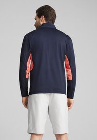 Herren Sportoberteil mit langen Ärmeln in Marineblau und pinken Seiteneinsätzen; leichtes, glattes Material. Getragen mit hellgrauen Shorts, von hinten stehend.