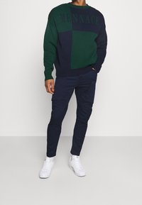 Grün und marineblau color-blocked Pullover mit "MENNACE"-Text, kombiniert mit marineblauen Cargo-Hosen und weißen Sneakers. Gestrickte Textur, lässige Passform.