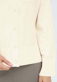 Kreminės spalvos kardiganas su raukšlėtu tekstūru, turintis penkis mygtukus ir prigludus�į hemą. Medžiaga atrodo minkšta ir šiek tiek elastinga.