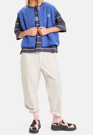 Personne portant une veste polaire bleue, un t-shirt oversize rayé noir et rose, un pantalon beige retroussé, des chaussettes roses et des chaussures marron et blanches sans lacets.