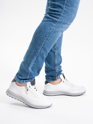 Weiße Leder-Sneaker mit Schnürsenkeln und seitlichem Reißverschluss, getragen zu blauen Slim-Fit-Jeans, an einer Person stehend vor weißem Hintergrund.