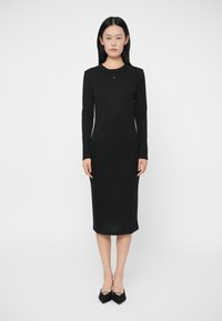 Γυναίκα που στέκεται μπροστά, φορώντας ένα εφαρμοστό μαύρο μακρυμάνικο φόρεμα midi και μαύρες μυτερές γόβες, μπροστά σε ουδέτερο λευκό φόντο.