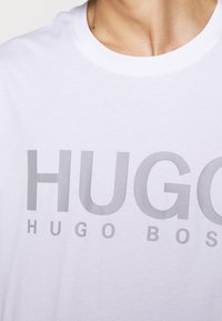Hvid bomulds t-shirt med stor lysegrå tekst "HUGO" og "HUGO BOSS" på tværs af brystet. Rund halsudskæring, glat tekstur.