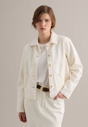 Giovane donna con capelli castani corti indossa una giacca avorio abbottonata sopra un top bianco e una gonna abbinata, mano in tasca, sfondo neutro.