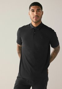 Polo nero con colletto, patta a tre bottoni, maniche corte e logo ricamato sul petto, abbinato a pantaloni scuri.