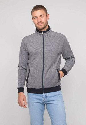 Homme portant une veste grise zippée avec des poignets et un col noirs, debout avec une main dans la poche et un jean bleu clair.