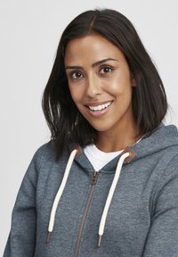Grå zip-up hoodie med vita dragkedjor och bruna accenter. Slät textur, med en frontdragkedja och en huva. Modellen har ett neutralt uttryck.