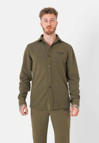 Camicia verde oliva a maniche lunghe con chiusura a bottoni, realizzata in tessuto morbido. Presenta un logo sul lato sinistro del petto e un design del colletto classico.