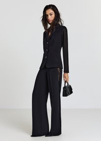 Blazer nero su misura con colletto a rever e chiusura con bottone, abbinato a pantaloni neri a gamba larga con pannelli laterali in pizzo, tenuto con una piccola borsa a mano.