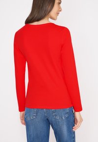 Camicia rossa a maniche lunghe con collo rotondo, realizzata in cotone liscio. La vista posteriore mostra un design aderente e cuciture minime. Abbinata a jeans in denim.