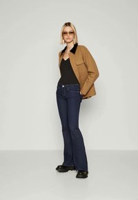Bruine jas met zwarte kraag, zwarte bodysuit, donkerblauwe flare jeans en chunky zwarte laarzen; staande model met recht blond haar.