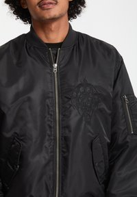 Bomber jacket nera realizzata in tessuto lucido, con zip frontale, colletto a coste e design ricamato sul petto. Tasche laterali incluse.