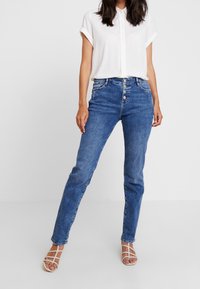 Jeans de mezclilla azul de talle alto con corte recto, cierre de cinco botones y presillas para cinturón; combinados con una camisa blanca con cuello y sandalias con tiras.
